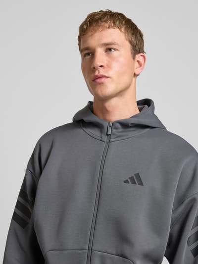 Männermodel trägt Sweatjacke mit Kapuze von adidas Sportswear in Grau, lockerer Schnitt, Reißverschluss, Label-Print, Nahaufnahme Oberkörper.