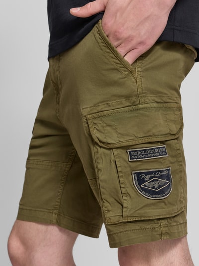 Mężczyzna nosi Regular Fit Bermudas mit Cargotaschen von Petrol, mieszanka bawełny z dodatkiem streczu, krój regular fit, wpuszczane kieszenie, naszywki z logo, kieszenie cargo, szerokość w rozmiarze M: 46 cm, długość wewnętrzna nogawki w rozmiarze M: 24 cm, oliwkowy kolor, tekstura gładka, detaliczne ujęcie nogawek.