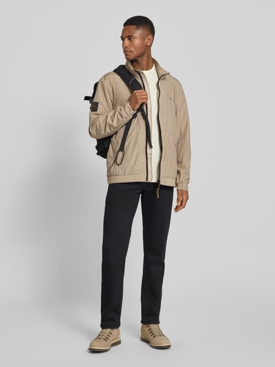 Männermodel trägt Blouson mit Logo-Print und Stehkragen von camel active in Beige, gerader Schnitt, Reißverschlusstaschen, Ganzkörperansicht von vorne.