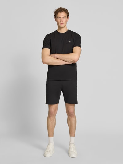 Männermodel trägt Regular Fit Sweatshorts aus reiner Baumwolle von Lacoste in Schwarz mit Logostreifen, Label Patch, elastischem Bund und 5-Pocket-Design, Ganzkörperansicht von vorne.