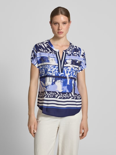 Frauenmodel trägt Blusenshirt aus Viskose mit Strasssteinbesatz von Betty Barclay, Statement-Motiv-Print, Tunikakragen, Kappärmel, Farbtöne Blau und Weiß, Straight Fit, Rückenlänge 55 cm, Ärmellänge 19 cm, Ganzkörperansicht von vorne.