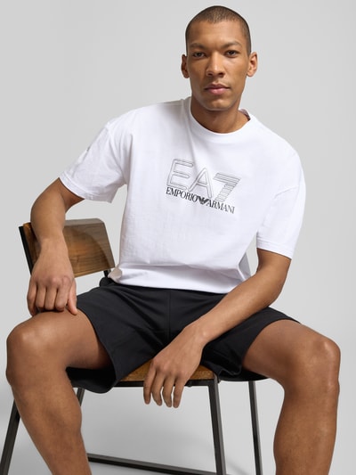 Męski model trägt T-shirt mit Logo und Rundhalsausschnitt von EA7 Emporio Armani z bawełny, krój regular fit, logo, okrągły dekolt, długość tyłu w rozmiarze M: 68 cm, ujęcie: całkowita przód.
