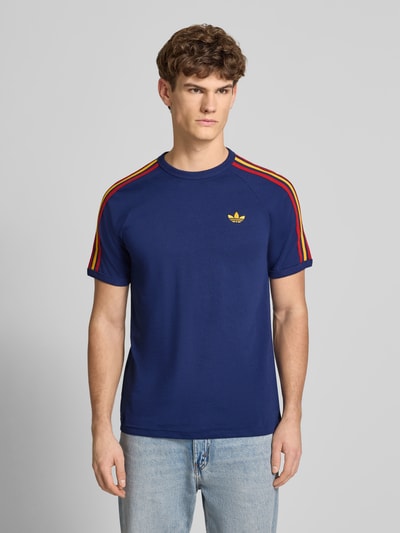 Männermodel trägt T-Shirt mit Label-Stitching von adidas Originals in Blau mit bunten Schulterstreifen, reine Baumwolle, krój regular fit, gerippte Abschlüsse, Rundhalsausschnitt, długość pleców w rozmiarze M: 74 cm, Ganzkörperansicht von vorne.