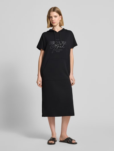 Frauenmodel trägt Sweatkleid mit Kapuze von Soyaconcept aus schwarzem Material-Mix mit Modal-Anteil, Label-Print, Gerader-Schnitt, Midikleid, Ganzkörperansicht von vorne.