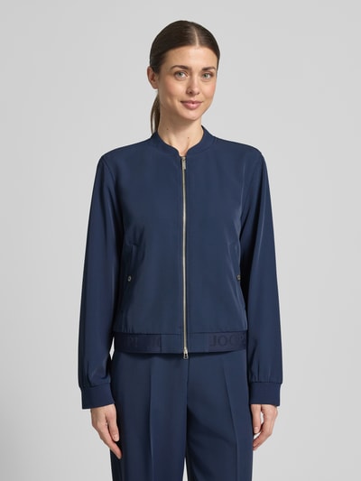 Kobiece model nosi kurtkę Jacke mit Zweiwege-Reißverschluss Modell 'Junipa' od JOOP!, mieszanka materiałów z dodatkiem streczu, krój regular fit, stójka, długość tyłu w rozmiarze 36: 52 cm, szerokość w rozmiarze 36: 52 cm, długość rękawów w rozmiarze 36: 62 cm, kolor granatowy, gładka faktura, zamek błyskawiczny dwustronny, widok całej sylwetki z przodu.