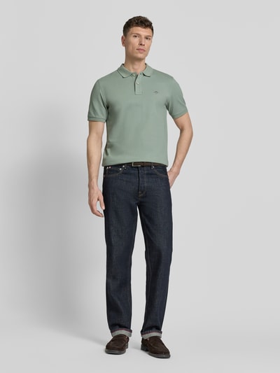 Męski model nosi Slim Fit Poloshirt mit Logo-Stitching od Gant z bawełny, krój slim fit, kołnierzyk polo, krótki rękaw, długość tyłu 70 cm w rozmiarze M, kolor zielony, gładka faktura, pełen widok z przodu.