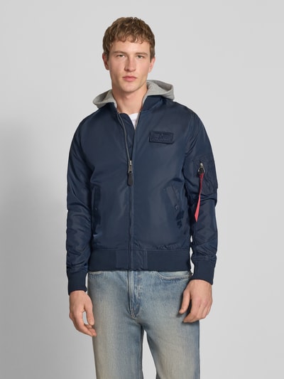 Männermodel trägt dunkelblauen Herren Blouson mit Logo-Anhänger Modell 'MA-1 TT' von Alpha Industries, Armtasche, Label-Patch, Ganzkörperansicht von vorne.
