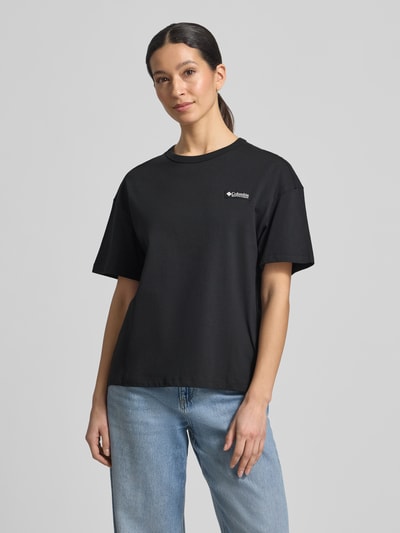 Frauenmodel trägt Damen T-Shirt aus reiner Baumwolle mit Modell 'CSC™' von COLUMBIA, lockere Passform, Rundhalsausschnitt, kurzärmelig, Label-Print, Omni-Shade Technologie UPF 30, Rückenlänge 60 cm bei Größe S, Schwarz, dezente Struktur, Ganzkörperansicht von vorne.