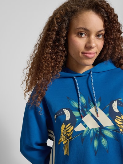 Modelka kobieca ma na sobie Regular Fit Hoodie aus Baumwoll-Mix od adidas Sportswear, efekt: jednolity kolor, nadruk z motywem, kaptur z tunelem, długi rękaw, prążkowane zakończenia, dzianina dresowa, mieszanka bawełny, zbliżenie na górną część ciała z przodu.
