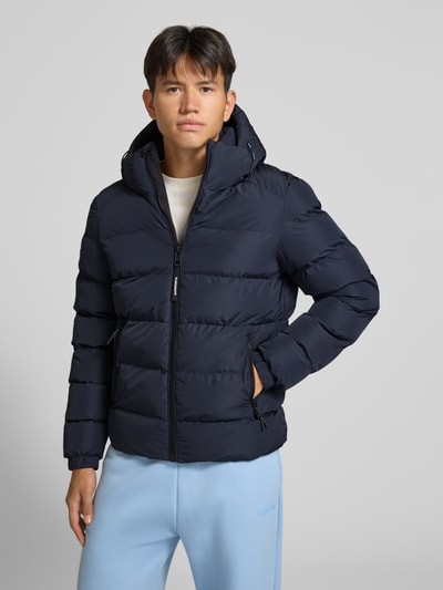Männermodel trägt Steppjacke mit Label-Stitching und Kapuze von Superdry in Dunkelblau, sichtbarer Reißverschluss, Ganzkörperansicht von vorne