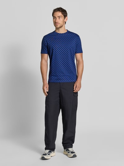 Męski model nosi Regular Fit T-Shirt mit Allover-Print od Armani Exchange w kolorze niebieskim z czarnym wzorem, okrągły dekolt, krój regular fit, nadruk na całej powierzchni, z krótkim rękawem, długość tyłu w rozmiarze M: 63 cm, Ganzkörperansicht von vorne.