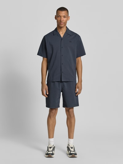 Mężczyzna nosi Relaxed Fit Freizeithemd aus reiner Baumwolle von Lacoste, tkanina stylizowana na denim, krój relaxed fit, kołnierzyk koszulowy, na całej długości listwa guzikowa, naszywka z logo, rękaw: krótki rękaw, wykończenie materiału: marszczenie, materiał: bawełna, kolor ciemnoniebieski, struktura crinkle, widok całej sylwetki z przodu.