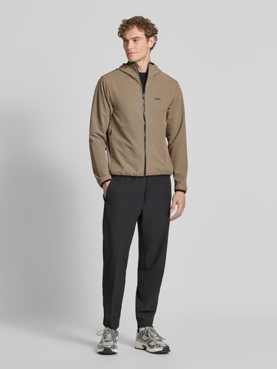 Männermodel trägt Regular fit trainingsjack met capuchon model 'LW_COMMUTERPRO' van BOSS Green in lichtbruin, zwarte broek, ganzkörperansicht van voren.