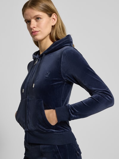 Frauenmodel trägt nachtblaue Sweatjacke mit Label-Stitching Modell 'ROBERTSON' von Juicy Couture, Material-Mix mit Stretch-Anteil, Regular-Fit, Kapuze mit Tunnelzug, Reißverschluss und Känguru-Tasche, samtige Textur, Nahaufnahme Oberkörper.