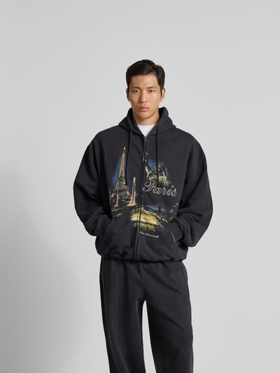 Männermodel trägt Oversized Sweatjacke mit Foto-Print von VETEMENTS aus Baumwoll-Mix, Oversized-Fit, Zweiwege-Reißverschluss, Kapuze mit Tunnelzug, dunkle Farben, weiche Textur, Rückenlänge 74 cm, Schulterbreite 68 cm, Ärmellänge 67 cm, Ganzkörperansicht von vorne.