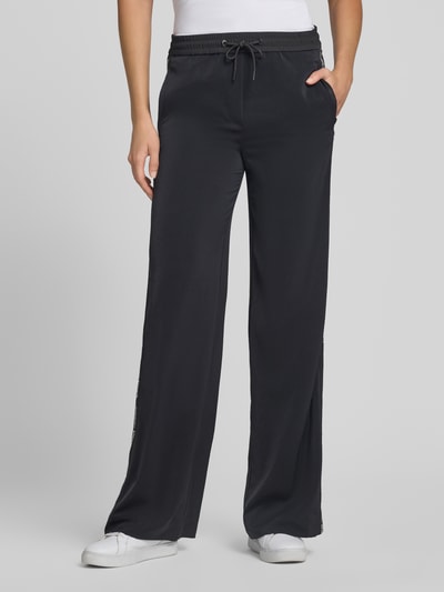 Frauenmodel trägt Schlupfhose mit seitlichen Label-Details und elastischem Bund von Marc Cain in Schwarz mit Eingrifftaschen, Relaxed-Fit, Ganzkörperansicht von vorne. Bundweite bei Größe 36: 39 cm, Innenbeinlänge bei Größe 36: 85 cm, Tunnelzug.