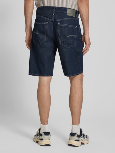 Männermodel trägt Regular Fit Jeansshorts aus reiner Baumwolle Modell 'DAKOTA SHORT' von G-Star in dunkelblauer Denim-Optik, 5-Pocket-Design, Label-Patch und Nieten, Materialverarbeitung Twill, Ganzkörperansicht Rückansicht.