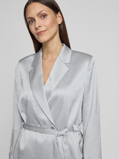 Frauenmodel trägt schimmernden silbergrauen Blazer mit Bindegürtel, glatte Textur, Nahaufnahme Oberkörper.