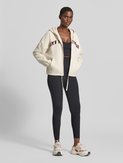 Frauenmodel trägt Sweatjacke mit Logo-Schriftzug von AIMN, cremefarben, Kapuze, Kängurutasche, Regular Fit, cropped, Ganzkörperansicht von vorne.