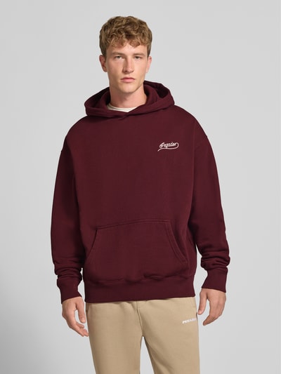 Männermodel trägt Regular Fit Hoodie aus Baumwoll-Mix von PEGADOR in Dunkelrot mit Kängurutasche und Strass-Logo, Ganzkörperansicht von vorne.