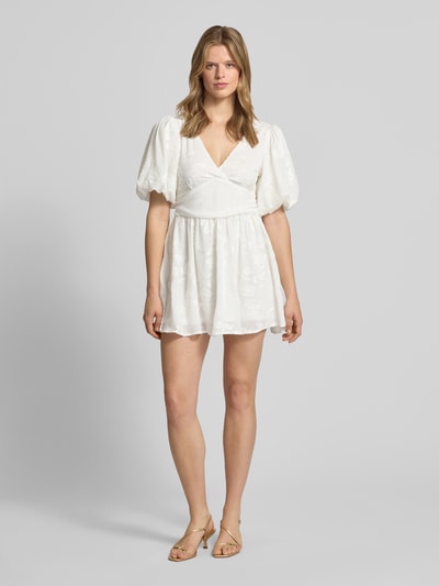 Frauenmodel trägt weißes Minikleid mit Ballonärmeln, tailliertem Schnitt, V-Ausschnitt, Embroidery Design von Gina Tricot, Ganzkörperansicht von vorne.