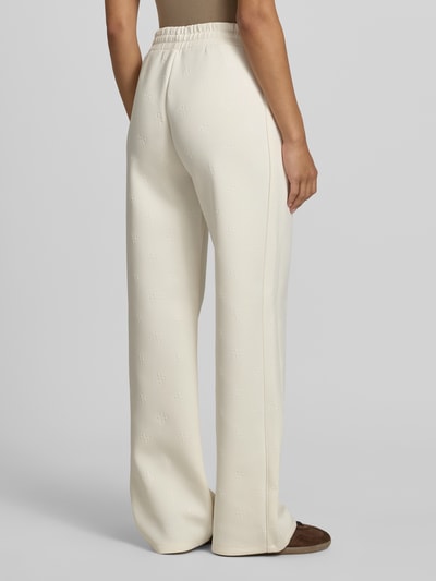 Frauenmodel trägt Wide Fit Sweatpants mit Allover-Logo-Print Modell 'Calista' von Guess Activewear in Beige, elastischer Bund, Ganzkörperansicht Rückansicht.