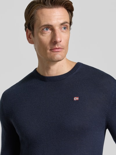 Męski model nosi sweter wełniany Slim Fit z czystej wełny Model 'DAMAVAND' od Napapijri, kolor granatowy, detal z logo, zbliżenie na górną część ciała, kontekst: sweter Napapijri krój slim fit z czystej wełny, detal z logo, elastyczne wykończenia, długość tyłu w rozm. M: 66 cm, długość rękawów w rozm. M: 69 cm.