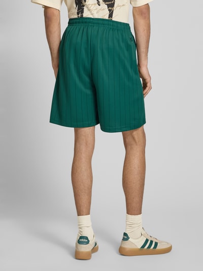 Männermodel trägt grüne Herren Shorts mit elastischem Bund, Modell 'Insignia Shadow' von KARL KANI im Allover-Look, Ganzkörperansicht Rückansicht. Bundweite 38 cm, Innenbeinlänge 19 cm bei Größe M.