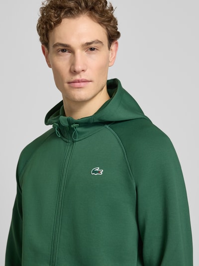 Męski model nosi Regular Fit Sweatjacke mit Baumwoll-Anteil od Lacoste Sport, bawełniany miks, logo, zamek, kaptur; zbliżenie na górną część ciała.