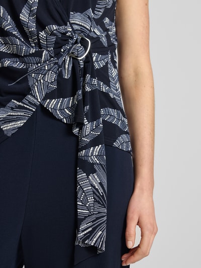 Kobieta nosi Relaxed Fit Jumpsuit in Wickel-Optik od Vera Mont z elastycznej mieszanki materiałów, kwiatowy nadruk, krótkie rękawy, marszczenia, okrągły dekolt, detal w stylu kopertowym, całkowita długość 150 cm, szerokość w rozmiarze 36: 36 cm, długość wewnętrzna nogawki 85 cm, ciemne kolory, proste linie, miękka tekstura, detal, zbliżenie górnej części.