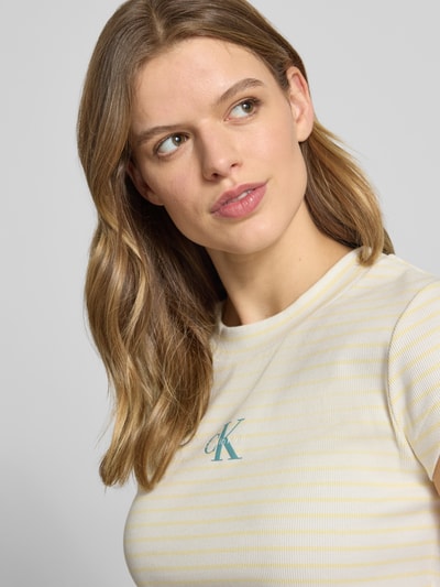 Modelka kobieca nosi Cropped T-Shirt mit Streifenmuster und Logo od Calvin Klein Jeans, bawełna z elastanem, wzór w paski, logo z przodu, dekolt okrągły, długość pleców w rozmiarze S: 39 cm, jasne kolory, tekstura prążkowana, zbliżenie na górną część ciała