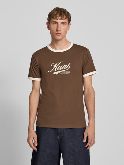 Męski model nosi T-Shirt mit Label-Print und Rundhalsausschnitt od Karl Kani w brązie z białymi detalami, bawełna, krój regular fit, długość tyłu w rozmiarze M: 68 cm, Ganzkörperansicht von vorne.