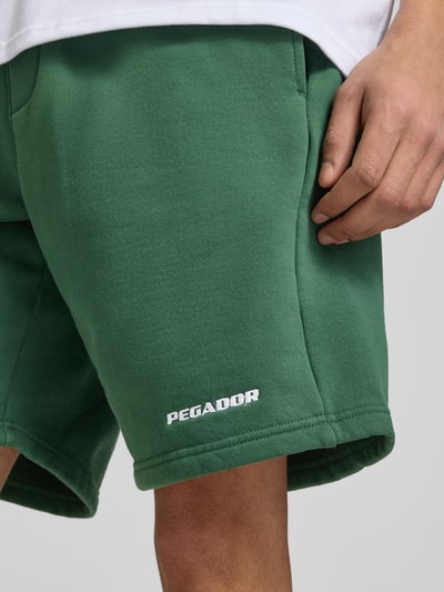 Mężczyzna nosi szorty mit elastischem Bund und Logo-Stitching von Pegador, mieszanka bawełny, krój loose fit, wpuszczane kieszenie, wyhaftowane logo, szerokość w rozmiarze M 38 cm, długość wewnętrzna nogawki 23 cm, zielony kolor, faktura gładka, detalaufnahme uda