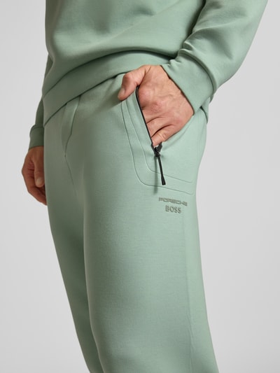 Männermodel trägt Regular Fit Sweatpants aus Baumwoll-Mix mit Modal-Anteil Modell 'PS SPIRIT70 HICON' von BOSS Green X Porsche, unifarbene Optik, seitliche Reißverschlusstasche, Label Print, Nahaufnahme Hüfte und Hand.