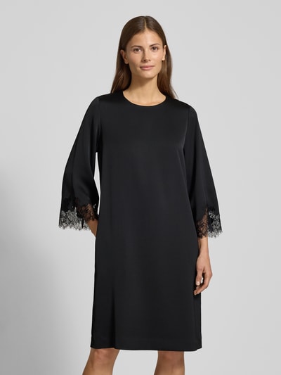 Kobiece model nosi sukienkę knielange Kleid mit Rundhalsausschnitt Modell 'Dancia' od JOOP!, krój relaxed fit, okrągły dekolt, długość do kolan, rękaw o dł. 3/4 z obszyciem koronką, długość tyłu 100 cm, szerokość klatki piersiowej 49 cm, długość rękawa 65 cm, gładka czarna tkanina, widok całej sylwetki z przodu.