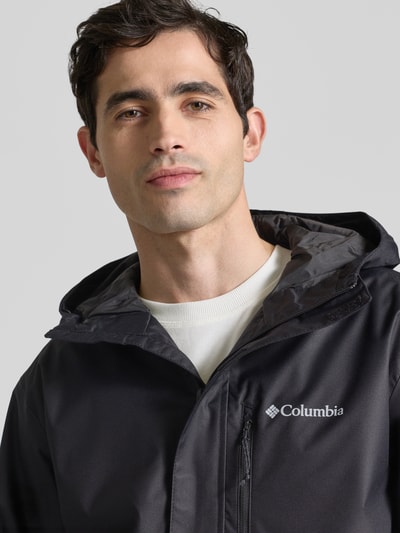 Męski model nosi Kurtka z kapturem Model 'Hikebound™ II' od Columbia, czarna, gładka faktura, logo, detal z zamkiem błyskawicznym, krój regular fit, kaptur z tunelem, patki przy mankietach, szerokość ramion w rozmiarze M: 49 cm, szerokość klatki piersiowej w rozmiarze M: 61 cm, długość tyłu w rozmiarze M: 74 cm, długość rękawów w rozmiarze M: 70 cm, zbliżenie na górną część ciała