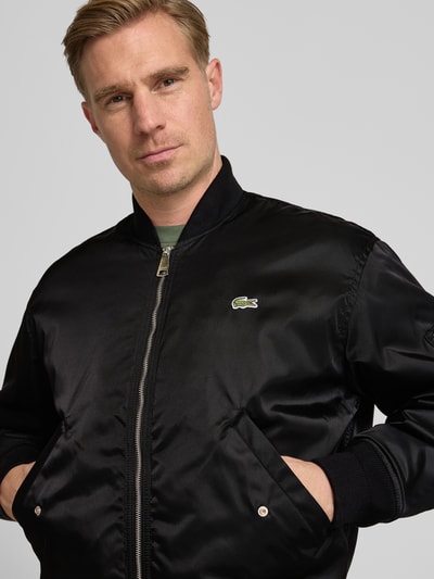 Männermodel trägt Regular Fit Bomberjacke aus Satin von Lacoste in Schwarz, unifarben, College-Kragen, Nahaufnahme Oberkörper.