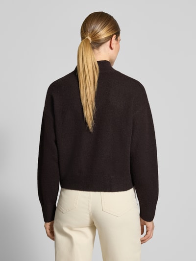 Frauenmodel trägt Turtleneck-Pullover mit Woll-Anteil und Rundhalsausschnitt von Neo Noir in Dunkelbraun, cremefarbene Hose, strukturierte Strickoptik, überschnittene Schultern, gerippte Abschlüsse, Ganzkörperansicht Rückansicht.