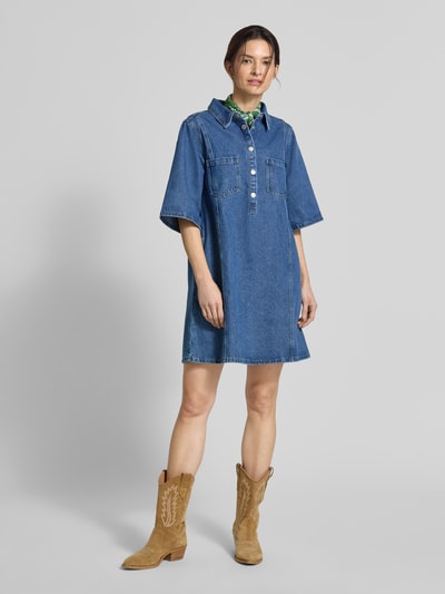 Frauenmodel trägt Jeanskleid mit Brusttaschen Modell 'Mola' von LOLLYS LAUNDRY aus Baumwoll-Viskose-Mix in Blau, Gerader-Schnitt, Knopfleiste, Hemdblusenkragen, Minikleid, Ganzkörperansicht von vorne.