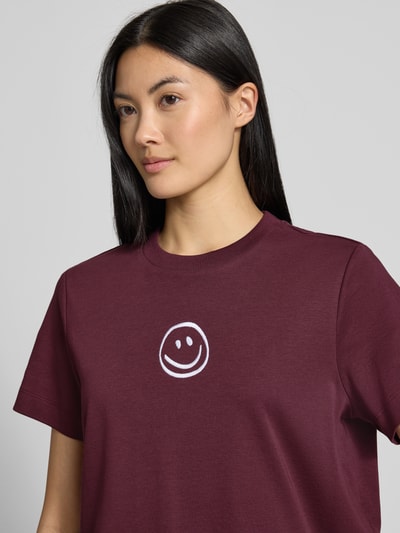 Kobiece modelka nosi damski t-shirt Boxy Fit T-Shirt aus reiner Baumwolle model 'SARA' od VERO MODA w kolorze bordowym z białym nadrukiem uśmiechu, gładka tekstura, zbliżenie na górną część ciała.