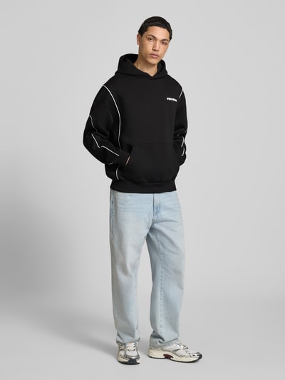 Męski model nosi Hoodie mit Logo und Känguru-Tasche od REVIEW, czarny, krój regular fit, bawełniany materiał, logo, kieszeń z przodu, długość tyłu w rozmiarze M: 71 cm, długość rękawa: 67 cm, pełny widok z przodu.