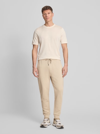 Männermodel trägt Regular Fit Sweatpants aus reiner Baumwolle von ARMANI EXCHANGE in Beige, mit elastischen Abschlüssen, Kordelzug, Seitentaschen, Gesäßtaschen, Label-Print, Innenbeinlänge 44 cm bei Größe M, Ganzkörperansicht von vorne.