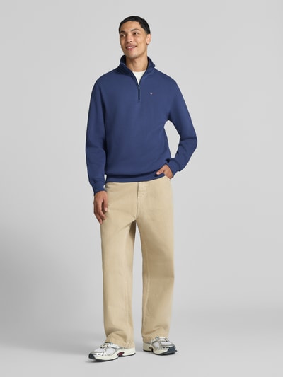 Männermodel trägt Regular Fit Sweatshirt aus Baumwoll-Mix von Tommy Jeans in Dunkelblau, helle Hose, weiße Sneaker, Ganzkörperansicht von vorne. Herren Sweatshirt von Tommy Jeans
