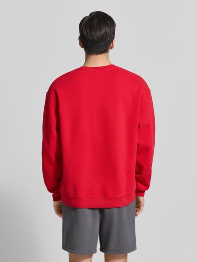 Męski model nosi bluzę Sweatshirt mit Logo Modell 'PLIZZY' od American Vintage, czerwień, bawełna, krój relaxed fit, okrągły dekolt, długość tyłu w rozmiarze M: 66 cm, długość rękawów w rozmiarze M: 60 cm, widok całej sylwetki od tyłu.
