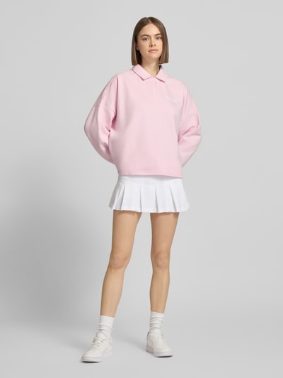 Frauenmodel trägt Sweatshirt mit Label-Print von adidas Sportswear in Hellrosa, weiter Schnitt, Hemdblusenkragen, Ganzkörperansicht von vorne.