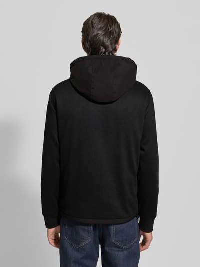 Männermodel trägt Windbreaker mit Zweiwege-Reißverschluss von Guess in Schwarz, Regular Fit, Kapuze, Rückenlänge 69 cm, Reißverschlusstaschen, Ganzkörperansicht Rückansicht.