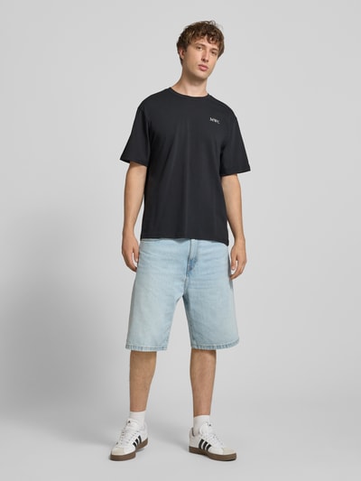 Mężczyzna model nosi Męski t-shirt Relaxed Fit T-Shirt aus reiner Baumwolle Modell 'CAP' von ONLY, czarny, z białym nadrukiem, jasnoniebieskie szorty, pełny widok z przodu.