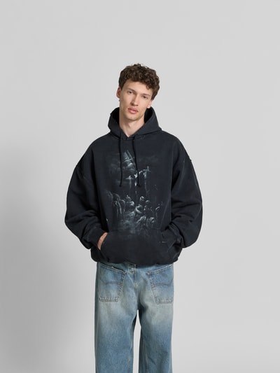 Männermodel trägt Oversized Hoodie aus Baumwoll-Mix von VETEMENTS in Schwarz mit Destroyed-Look, Motiv-Print, Känguru-Tasche, Ganzkörperansicht von vorne. Rückenlänge bei Größe M: 76 cm.