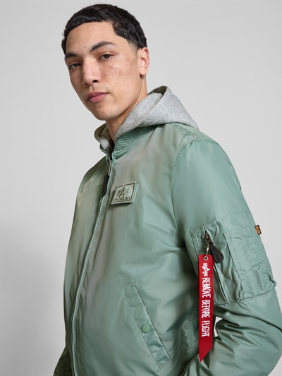 Männermodel trägt mintgrünen Blouson mit Logo-Anhänger Modell 'MA-1 TT' von Alpha Industries, graue capuchon van sweatstof, labelpatch, mouwzak met logohanger, los model, elastische boordjes, ritssluiting, ruglengte maat M 69 cm, schouderbreedte 46 cm, borstwijdte 54 cm, mouwlengte 67 cm, Nahaufnahme Oberkörper.