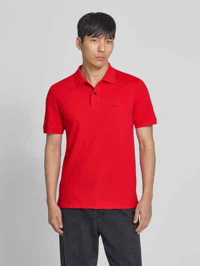 Männermodel trägt Regular Fit Poloshirt aus reiner Baumwolle Modell 'PALLAS' von BOSS, Farbe Rot, Optik: Unifarben, Piqué-Struktur, Label-Stitching-Applikation, Polokragen, V-Ausschnitt, kurze Knopfleiste, Kurzarm, gerippter Ärmelabschluss, Rumpfabschluss mit Seitenschlitzen, Ganzkörperansicht von vorne.